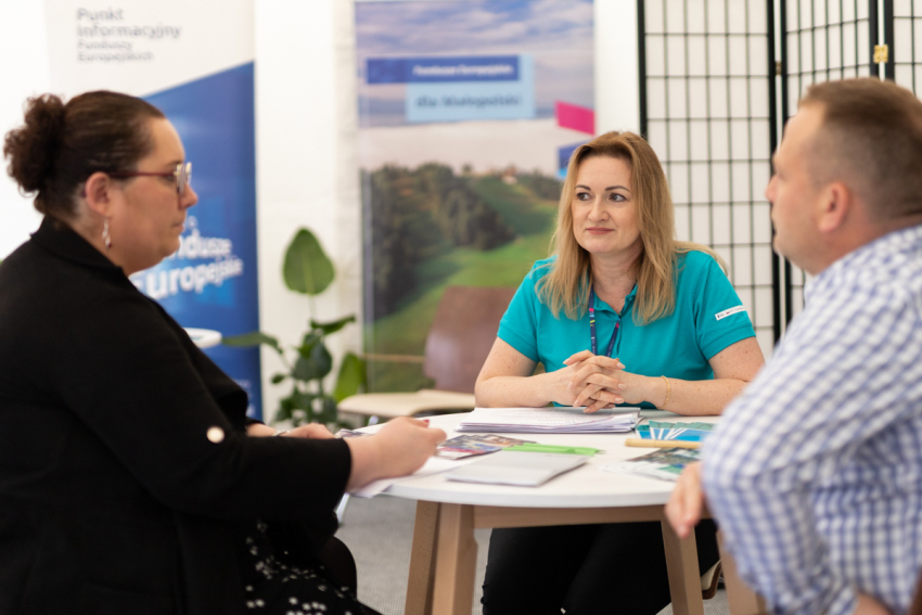 Na pierwszym planie troje ludzi siedzi przy okrągłym stole, na którym znajdują się ulotki i notesy. Na drugim planie dwa roll-upy.