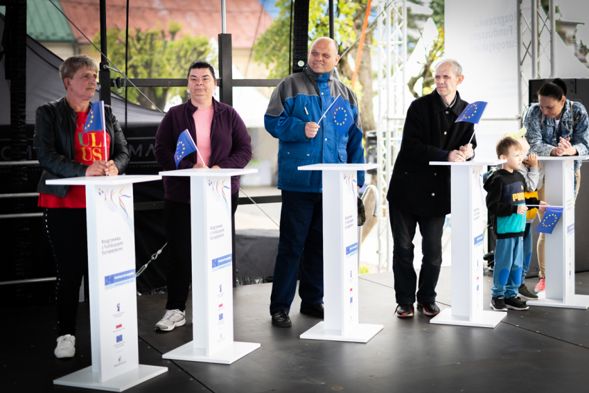 Piątka dorosłych osób stoi przy białych mównicach. W ręku trzymają chorągiewki z flagą Unii Europejskiej.