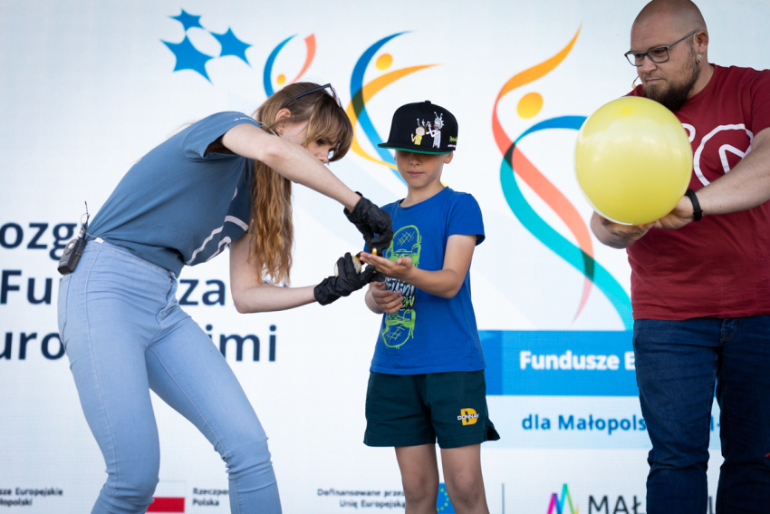 Po prawej mężczyzna trzymający balon, po lewej pochylona kobieta w czarnych rękawiczkach wręcza chłopcu jakiś przedmiot. W tle ścianka reklamowa „Fundusze Europejskie dla Małopolski”.