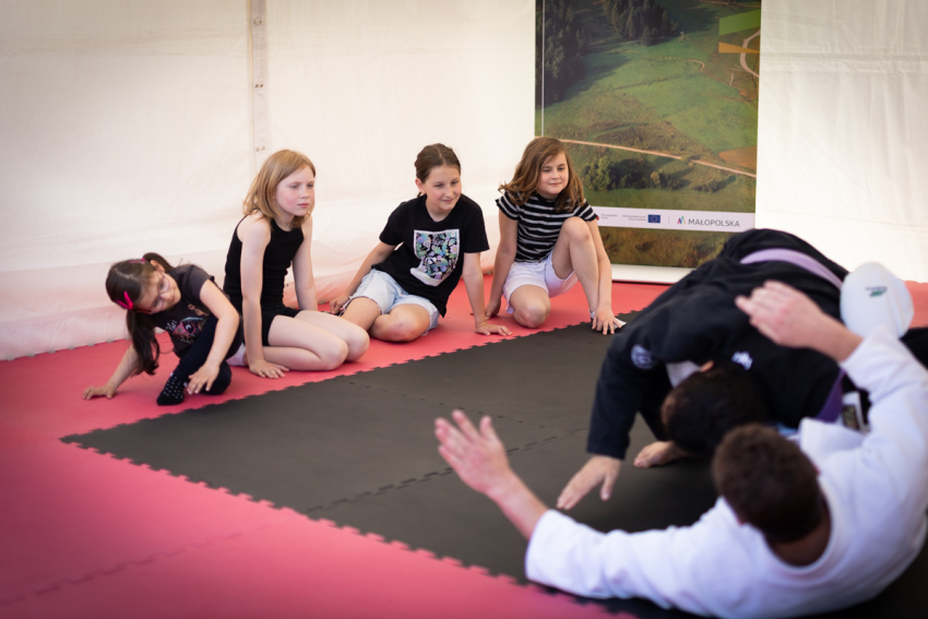 Dwóch mężczyzn w kimono tarza się po macie. Obok cztery dziewczynki siedzą i przyglądają się im z zaciekawieniem.