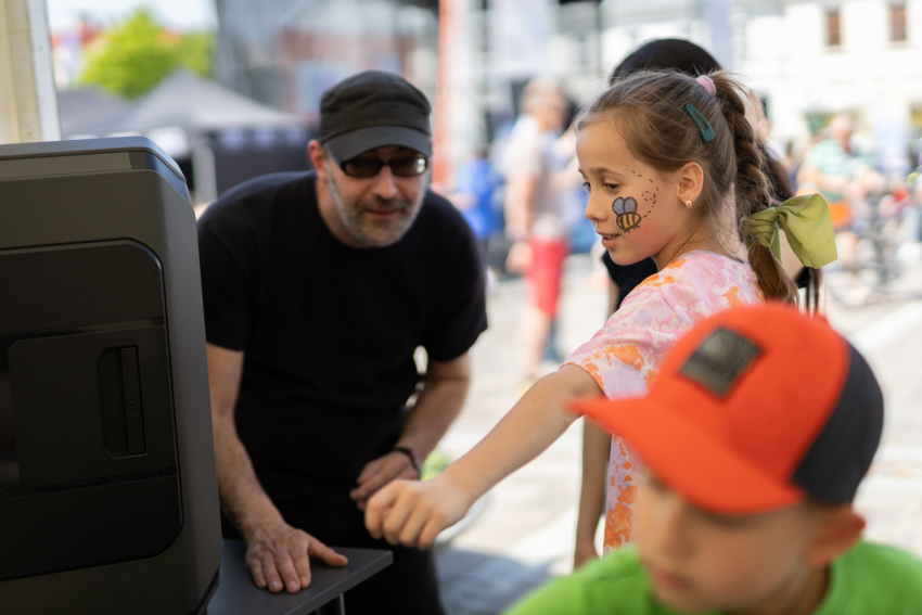 Dziewczynka z pomalowaną twarzą zerka do środka drukarki 3D. Obok mężczyzna i kilkuletni chłopiec – obaj w czapkach z daszkiem.