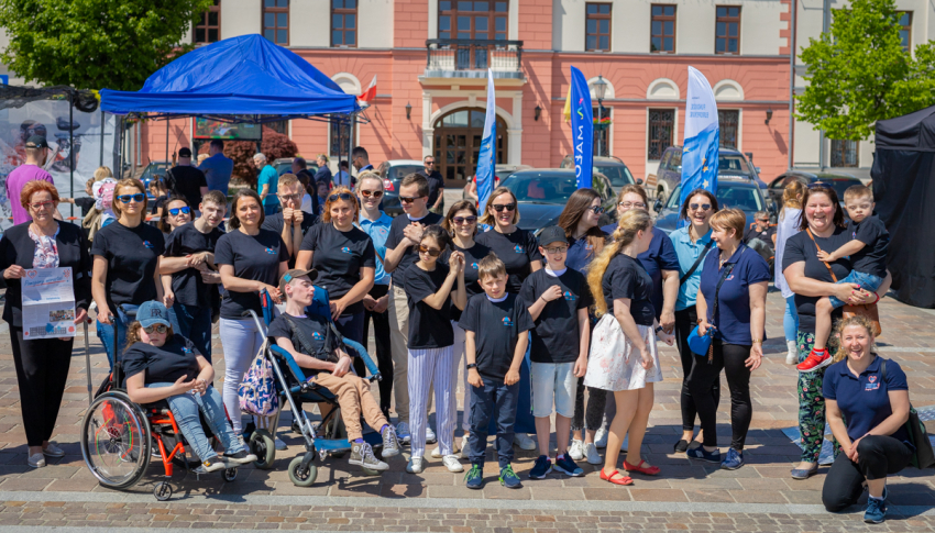 Kilkanaście osób w jednakowych koszulkach pozuje do zdjęcia. Wśród nich dwie osoby na wózkach inwalidzkich. W tle flagi reklamowe i namiot.