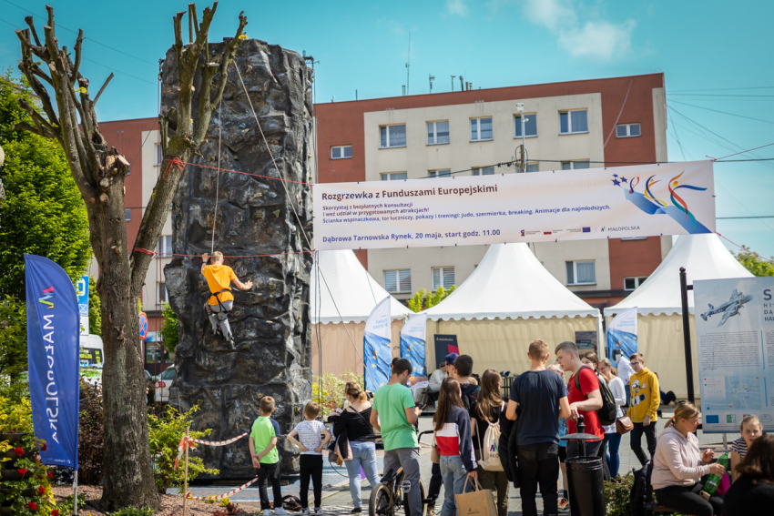 Na pierwszym planie grupa ludzi i flaga reklamowa z napisem „Małopolska”. Na dalszym planie ścianka wspinaczkowa, z wspinającym się na nią mężczyzną. W głębi namioty.