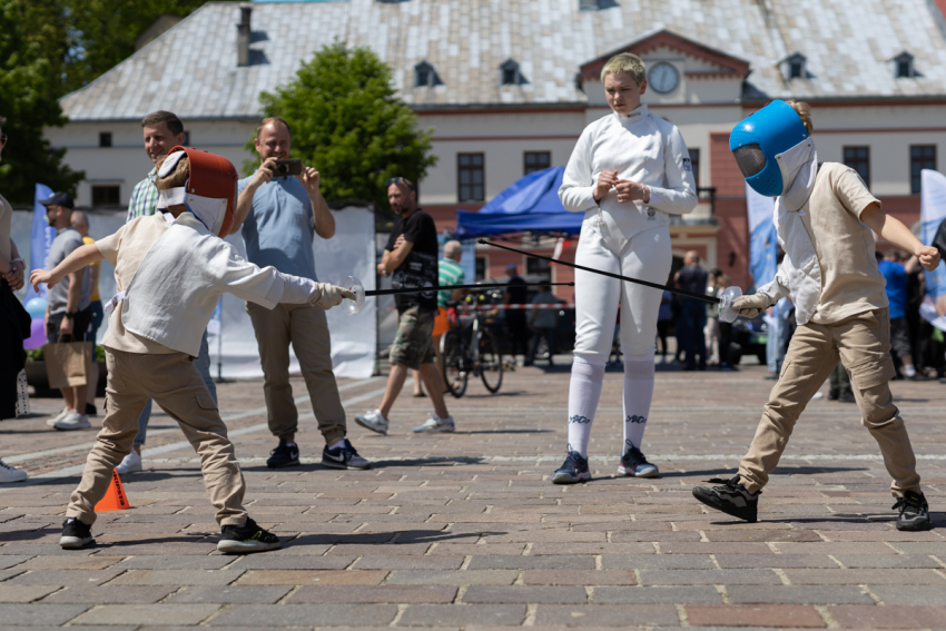 Dwóch chłopców w kaskach walczy drewnianymi kijami. Obok stoi kobieta w stroju szermierza i mężczyzna, który robi zdjęcie telefonem. W tle zabytkowy budynek.