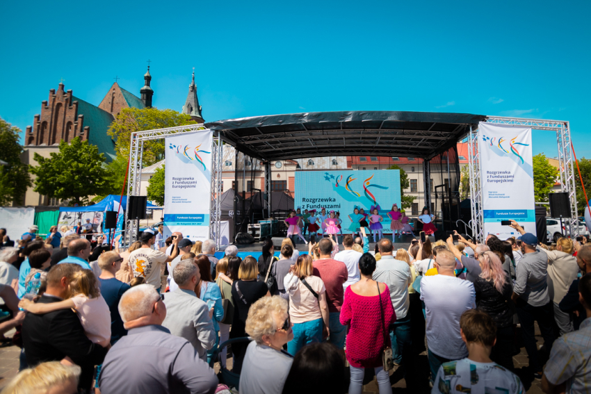 Na pierwszym planie tłum ludzi. Na drugim planie zadaszona scena z dużym ekranem projekcyjnym. Na scenie występują dzieci. W tle kościół oraz bezchmurne niebo.