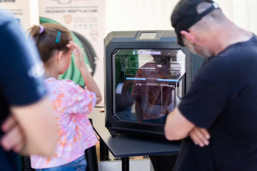 Dziewczynka i mężczyzna spoglądają do środka drukarki 3D. W głębi dwa roll-upy.