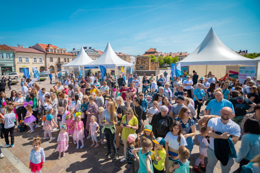 Na pierwszym planie łum ludzi w różnym wieku. Na drugim planie namioty imprezowe ze spiczastym zadaszeniem. W tle kamienice i błękitne niebo.