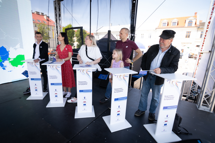 Pięć dorosłych osób stoi przy białych mównicach. Obok jednej z nich stoi kilkuletnia dziewczynka.