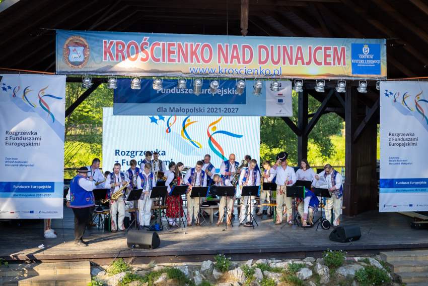 Zespół muzyczny w ludowych strojach góralskich na scenie, która  znajduje się w plenerze. Na scenę pada światło słoneczne, w tle widoczne drzewa. Po obu stronach sceny oraz na środku , za zespołem informacje na temat  Rozgrzewki z Funduszami Europejskimi. Na górze wisi duży napis &quot;Krościenko Nad Dunajcem&quot;.