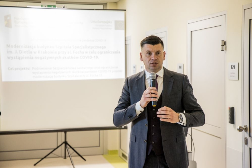 Młody mężczyzna stoi i trzyma w ręku mikrofon. Za nim, na rozłożonym ekranie wyświetlane są informacje na temat projektu modernizacji szpitala.