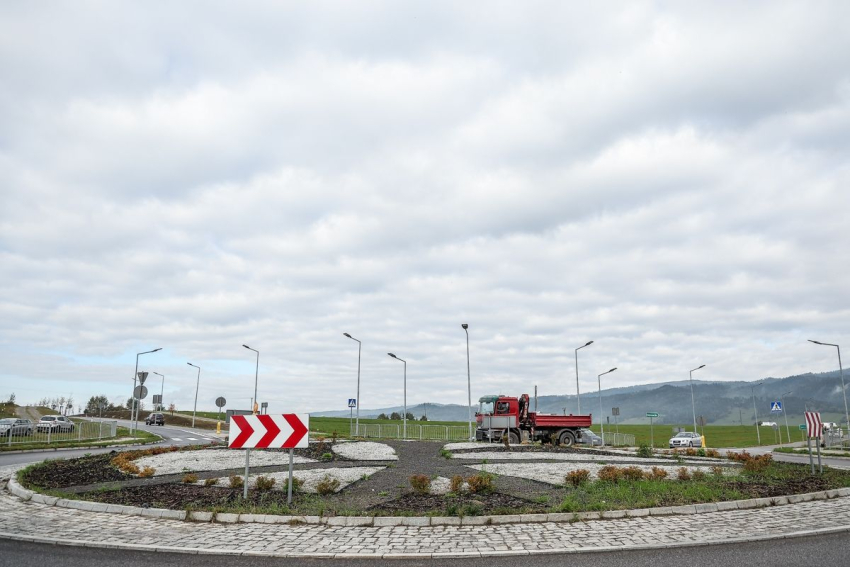 Rondo. Od prawej strony wjeżdżająca czerwona ciężarówka, samochód osobowy. W oddali zielona trawa, wzgórza i niebo usłane chmurami.