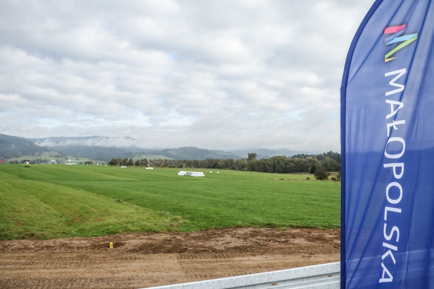 Rozległa, zielona polana. Z przodu zdjęcia widoczny fragment bariery zabezpieczającej obok drogi. Z prawej strony fragment windera/flagi z napisem Małopolska. W oddali zielone wzgórz i unosząca się nad nimi mgła.