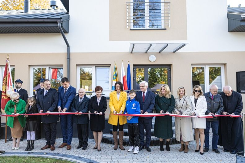 Kilkanaście osób w różnym wieku stoją przed nowym budynkiem i przecinają wstęgę na znak otwarcia .