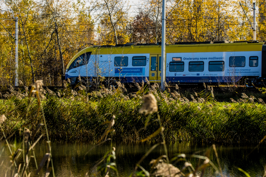 Żółto, biało, niebieski pociąg z napisami : Otwieramy nowe możliwości, Fundusze Europejskie. Pociąg przejeżdża przez las lub park -drzewa, trawy, widoczny fragment rzeki lub stawu. Słoneczny dzień.