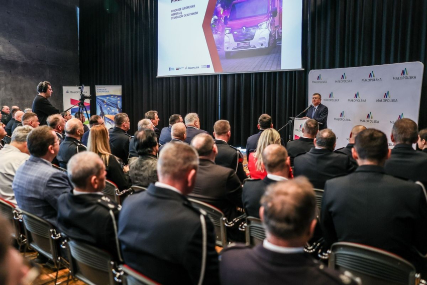 Marszałek województwa małopolskiego przemawia do zebranych ludzi w sali konferencyjnej. Za Panem Marszałkiem na ekranie projektora widoczny samochód strażacki. Obok duża plansza z logo Małopolska.