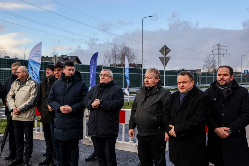 Kilku mężczyzn ubranych w zimowe kurtkii płaszcze stoi wzdłuż drogi. Za nimi widoczny fragment barierki drogowej oraz windery z logo Małopolski i Funduszy Europejskich. 