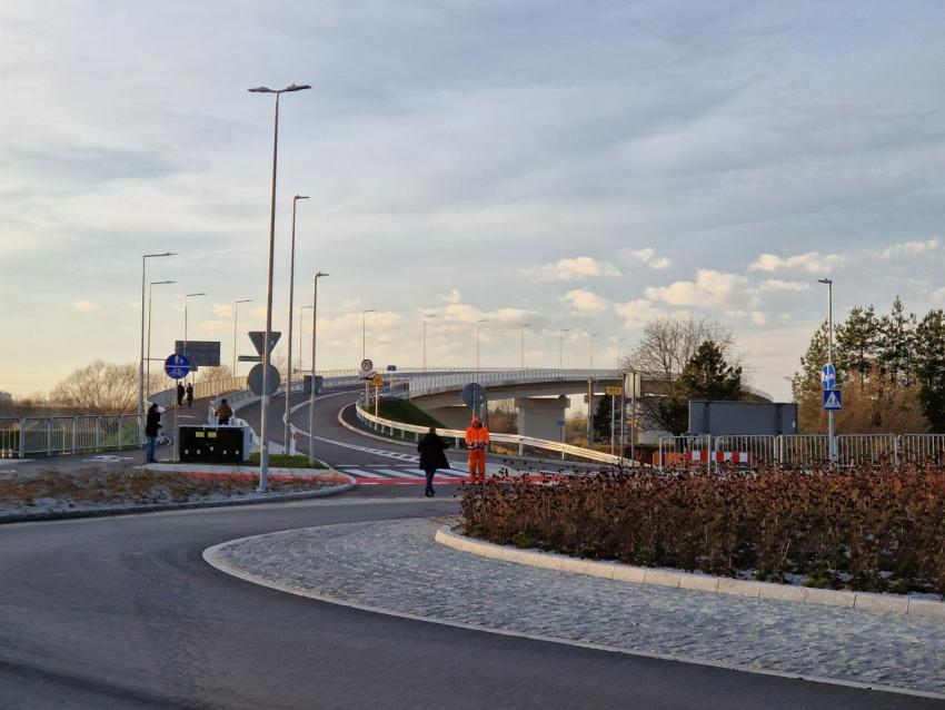 Rondo. Pośrodku wysepka z suchymi kwiatami. Od ronda odchodzi most. Przejście dla pieszych, ścieżka rowerowa. Po lwesj stronie mostu 4 osoby, spacerują, jadą na rowerze. Przy ścieżce rowerowej stoi mężczyzna w pomarańczowym kombinezonie roboczym i drugi w ciemnej kurtce.