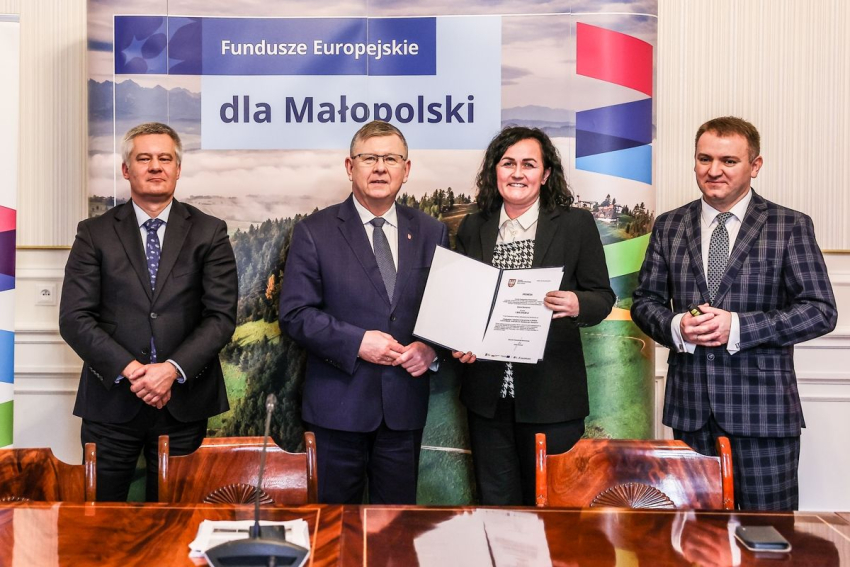 Trzech mężczyzn i jedna kobieta stoją przy stole konferencyjnym. Wszyscy są elegancko ubrani i lekko uśmiechnięci. Kobieta trzyma w ręku dokument, który prezentuje do zdjęcia. Za nimi plakat z napisem Fundusze Europejskie dla Małopolski.