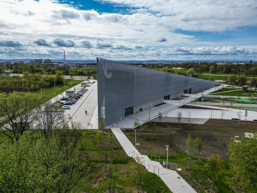 Nowoczesny budynek Centrum Nauki Cogiteon. Po lewej stronie parking samochodowy, na środku ścieżka wyłozona kostką brukową, wokół zieleń-trawa, krzewy i drzewka. W tle błękitne niebo usłane chmurami.