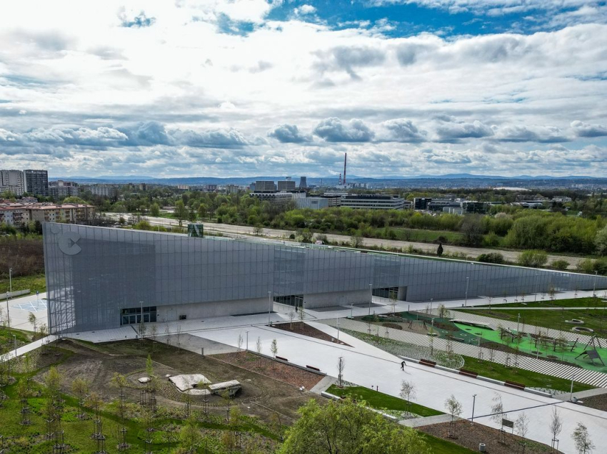 Bardzo nowoczesny budynek Cnetrum Nauki Cogiteon. Budynek ma bryłę trójkąta i jest przeszklony. Wokoł budynku ściezki dochodzące i park. W oddali widoczne inne nowoczesne budynki i kominy fabryk. Niebo usłane chmurami.