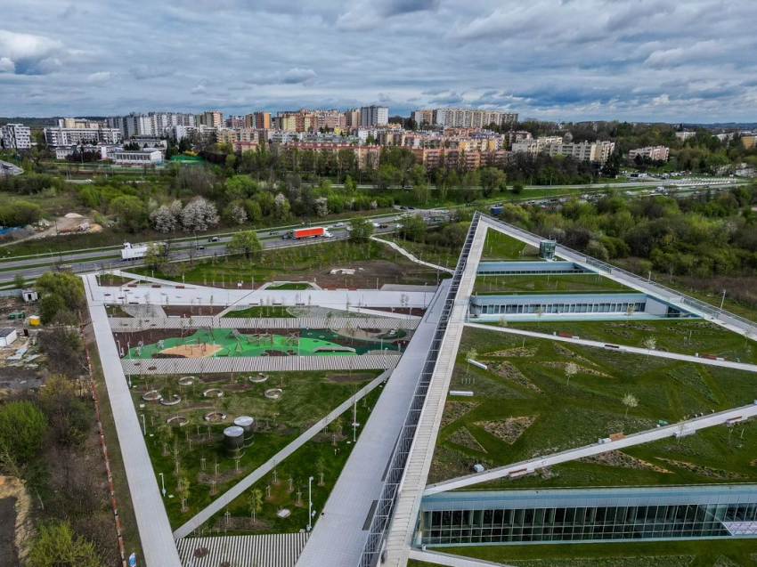 Budynek Centrum Nauki oraz przestrzeń wokół widziana z góry. W oddali osiedle mieszkalne, bloki, park, droga z jadącymi samochodami.