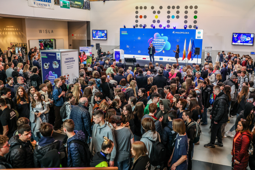 Tłum ludzi w dużej hali. Pod ścianą scena, na której stoją dwie kobiety. Jedna z nich przemawia do mikrofonu. Za nimi  duża niebieska tablica z napisami : Festiwal Zawodów w Małopolsce 2024. Na dole pasek z logotypami Funduszy Europejskich, Małopolski, Rzeczposoplitej oraz Unii Europejskiej.