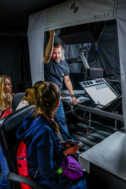 Młody mężczyzna w pracowni digitalizacji pokazuje kilku osobom sprzęt do digitalizacji.