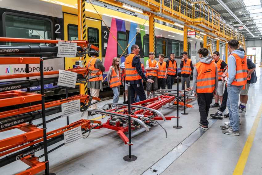 Szkolna grupa z opiekunką i mężczyzna z kolei, w pomieszczeniu obsługi taboru kolejowego. Po lewej stronie pociąg.