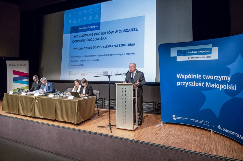 Scenka, na której przemawia mężczyzna przy mównicy. Obok stół przy, którym siedzą 3 osoby.