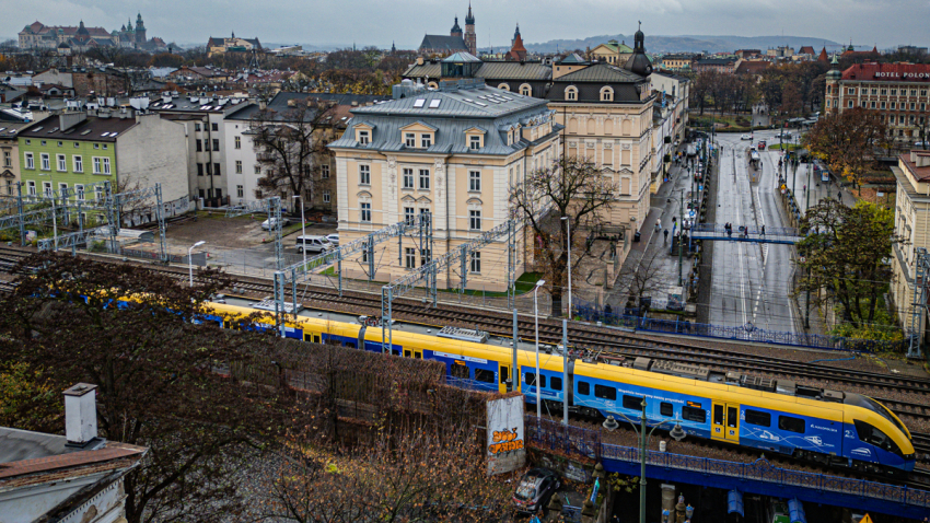 Pociąg przejeżdżający przez miasto. Kamienice, ulice, ludzie, drzewa. Widok z lotu ptaka.