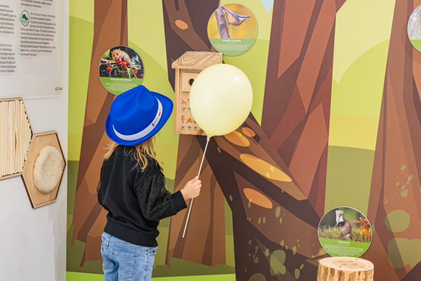 Dziewczynka w kapeluszu i z balonikiem w ręku stoi przy karmniku dla ptaków.