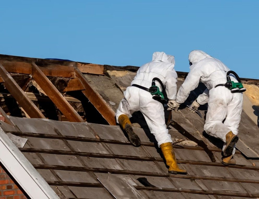 Dwóch ludzi w kombinezonach ściąga azbest z dachu.