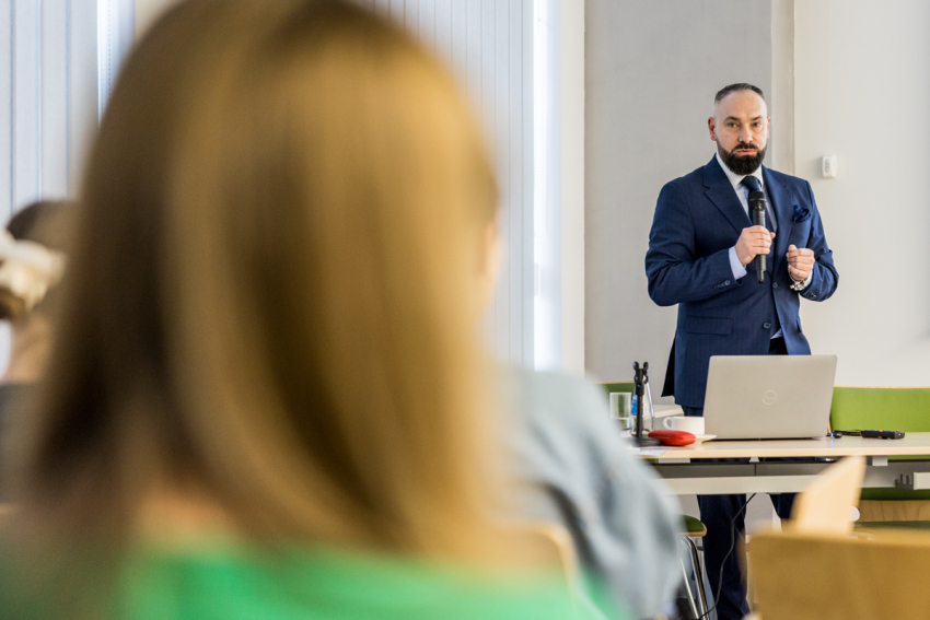 Na pierwszym planie ujęcie od tyłu grupę osób siedzących na krzesłach w sali szkolenowej. W głębi kadru prelegent trzymający w dłoni mikrofon