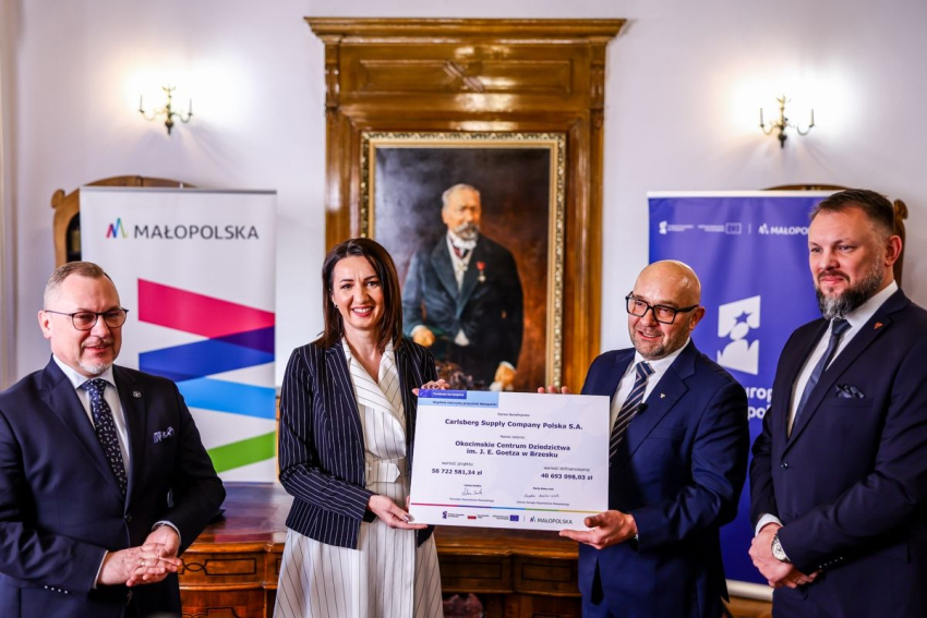 Grupa czterech elegancko ubranych osób stoi w reprezentacyjnym wnętrzu, prezentując duży certyfikat wsparcia finansowego dla Carlsberg Supply Company Polska S.A. W tle widoczne są banery promocyjne Małopolski i Funduszy Europejskich oraz portret historycznej postaci na ścianie.