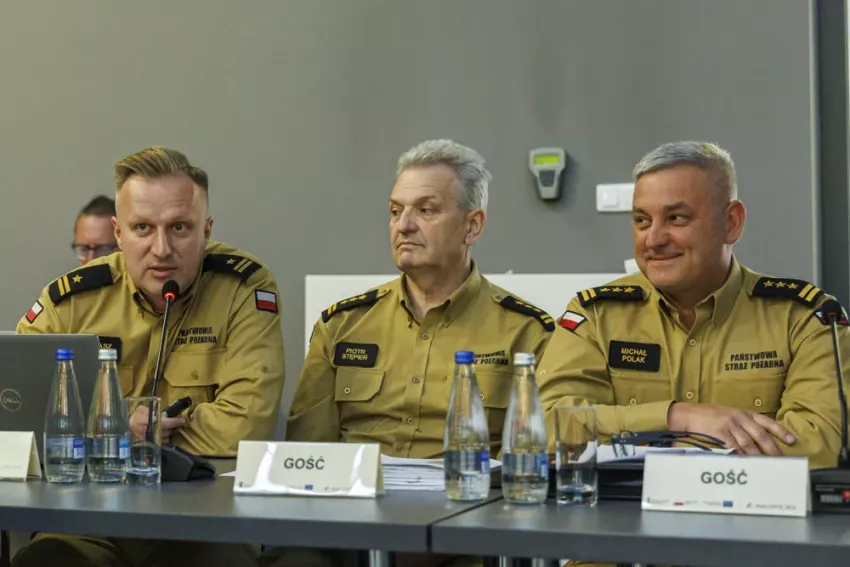 Trzech funkcjonariuszy Państwowej Straży Pożarnej siedzących przy stole konferencyjnym, z których jeden przemawia do mikrofonu