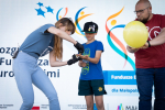 Po prawej mężczyzna trzymający balon, po lewej pochylona kobieta w czarnych rękawiczkach wręcza chłopcu jakiś przedmiot. W tle ścianka reklamowa „Fundusze Europejskie dla Małopolski”.