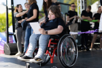 Kobieta siedzi na wózku inwalidzkim. W jednej ręce trzyma kartkę, w drugiej mikrofon. W tle grupa młodych ludzi.
