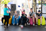 Pięć młodych osób siedzi na różnokolorowych pufach. Trzymają chorągiewki z flagami Unii Europejskiej. Obok stoi kobieta z mikrofonem.