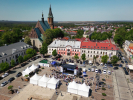 Widok z lotu ptaka na rynek miejski otoczony kamienicami. W centrum ustawione białe namioty imprezowe oraz scena z zadaszeniem. Przed sceną tłum ludzi, a za sceną zaparkowane samochody.