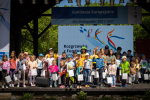 Duża grupa dzieci w wieku przedszkolnym i wczesnoszkolnym stojąca na scenie, która znajduje się na zewnątrz , w otoczeniu drzew. W tle grafika z hasłami dotyczącymi Funduszy Europejskich.
