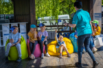 Grupka dzieci siedzących na kolorowych pufach , na scenie. W dłoniach mają niebieskie wiatraczki w żółte gwiazdki (barwy Unii Europejskiej). Przodem do dzieci stoi konsultantka Funduszy Europejskich z mikrofonem w lewej ręce. W prawej trzyma papierową torebkę na prezenty lub nagrody. Za sceną widoczne drzewa.