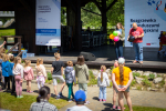 Grupka dzieci stojąca przed sceną w słoneczny dzień. Na scenie młoda kobieta i młody mężczyzna trzymający kolorowe balony. Za nimi widoczna informacja na temat odbywającego się wydarzenia czyli Rozgrzewki Z Funduszami Europejskimi.