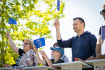 Na pierwszym planie starsza kobieta w ciemnych okularach, chłopiec w okularach i czapeczce z daszkiem oraz młody, uśmiechnięty mężczyzna. W rękach mają chorągiewki w kolorze granatowym z żółtymi gwiazdkami (barwy Unii Europejskiej). Po lewej stronie rozmazana korona drzew, przez którą przebija się słońce.