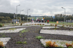 Na pierwszym planie fragment ronda, wysypane żwirek, kamyczki, kępki trawy. Na dalszym planie widoczna droga i malutkie postacie, kilkanaście osób , wóz strażacki. Po obu stronach drogi znaki drogowe, polanki i las. W oddali widoczne wierzchołki gór. Zachmurzone niebo.