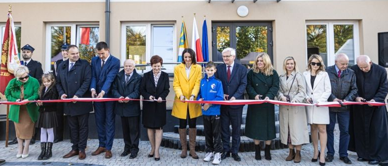 Kilkanaście osób w różnym wieku stoją przed nowym budynkiem i przecinają wstęgę na znak otwarcia .