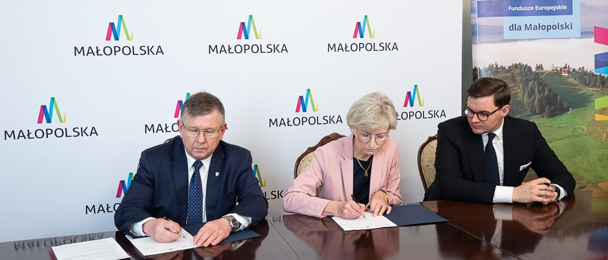 Kobieta i dwóch mężczyzn siedzą przy stole konferencyjnym. Jeden z mężczyzn i kobieta podpisują umowę. Wszyscy elegancko ubrani. Za nimi duża, biała plansza z napisami Małopolska. Obok planszy winder, plansza z napisem Fundusze Europejskie dla Małopolski.