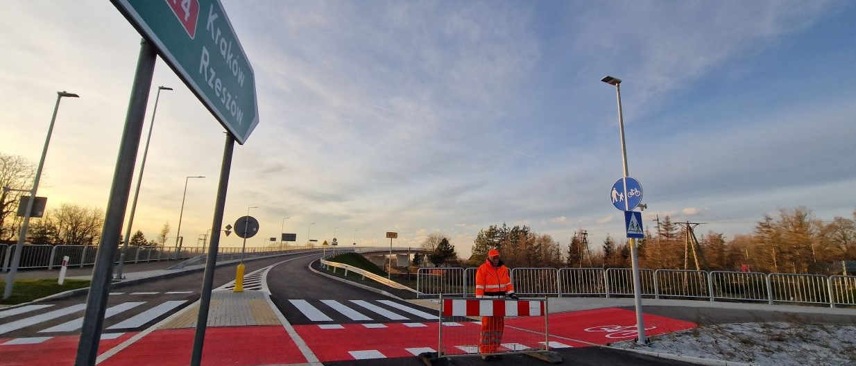 Most. Przejście dla pieszych, ścieżka rowerowa w kolorze czerwonym, pośrodku wysepka z kostki brukowej a na niej drogowskaz A4 Kraków-Rzeszów. Po prawej stronie, przy ścieżce rowerowej stoi mężczyzna ,ubrany w roboczy ,pomarańczowy kombinezon i przestawia płotek drogowy. W oddali , po obu stronach drzewa, latarnie, znaki drogowe. 