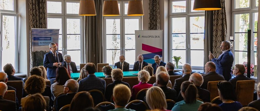 Zdjęcie przedstawia grupę osób w różnym wieku na spotkaniu w sali z dużymi oknami