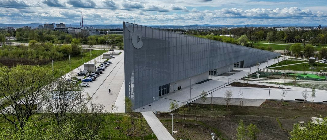 Nowoczesny budynek Centrum Nauki Cogiteon. Przed budynkiem parking samochodowy. Wokół budynku park.