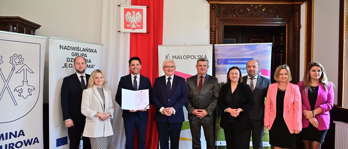 Grupka ludzi, 4 kobiety i 5 mężczyzn stoi w eleganckiej sali. Jeden z mężczyzn trzyma promesę.